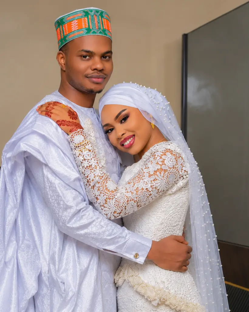 Jeunes mariés ouest-africains en tenue traditionnelle blanche, l’homme coiffé d’un bonnet brodé vert et orange, la mariée en voile de perles, souriant sous une arche violette.